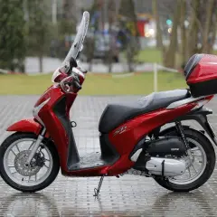 Czerwony skuter Honda SH z kufrem, gotowy do jazdy. W Polsce od ilu lat można jeździć skuterem? Od 14 lat na 50cc, od 18 lat na większe pojemności.