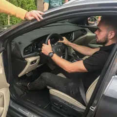 Mężczyzna siedzi za kierownicą, pokazując, jak ustawić fotel kierowcy. Dwie osoby pomagają mu, jedna trzyma telefon, druga opiera się o drzwi.
