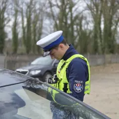 Policjant w kamizelce odblaskowej i czapce, pochylony nad maską samochodu, stosuje zasadę ograniczonego zaufania.
