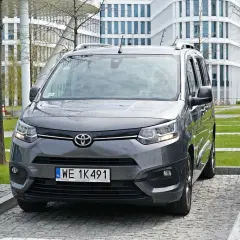 Szary Toyota Proace City zaparkowany prostopadle tyłem na parkingu przed nowoczesnym budynkiem.