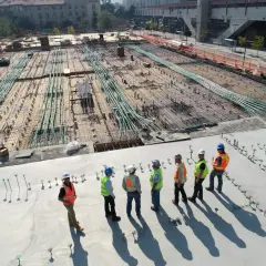 Grupa budowlańców w kaskach i kamizelkach obserwuje postęp prac na budowie, gdzie przygotowywana jest płyta fundamentowa zgodnie z art. 62 prawa budowlanego.