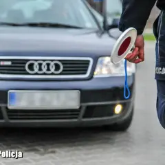 Policjant z lizakiem zatrzymuje auto. Widać jego mundur i fragment ciemnego Audi. Jakie światła podczas deszczu, gdy policja kontroluje ruch.