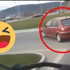 Czerwony samochód próbuje zawrócić na rondzie, ale robi to na lewo, co wygląda na niezbyt przemyślane. Obok śmieje się emotikon.