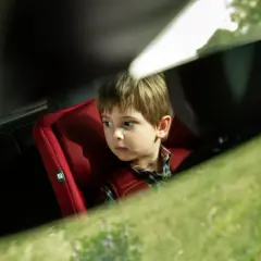 Zamyślone dziecko w aucie, siedzi w czerwonym foteliku. Za oknem zielona trawa i drzewa.
