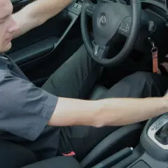 Mężczyzna w czarnej koszuli i spodniach, siedząc za kierownicą samochodu z automatyczną skrzynią biegów, czeka na rozwinięcie się korka.