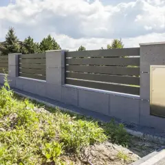 Nowoczesne ogrodzenie z betonowych słupków i ciemnych paneli, wyznaczające nieprzekraczalną linię zabudowy. Złota skrzynka na listy wmurowana w słupek.