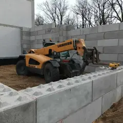Żółty ładowacz teleskopowy Liebherr układa betonowe bloki, budując mur oporowy.