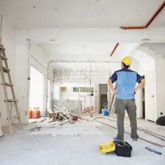 Pracownik w żółtym kasku nadzoruje remont. W pomieszczeniu widać drabiny, gruz i narzędzia. Prawo budowlane wymaga staranności.