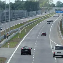 Samochody poruszają się lewym pasem w terenie zabudowanym, mijając ekrany akustyczne.