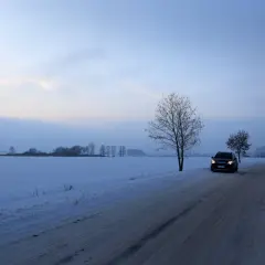 Samochód jedzie po ośnieżonej drodze wśród pól. Wczesny wieczór, niebo lekko zaróżowione. Jazda po śniegu wymaga ostrożności.