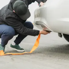 Mężczyzna mocuje pomarańczową linę holowniczą do samochodu. Pamiętaj o ograniczeniach prędkości, z jaką można holować samochód, by zapewnić bezpieczeństwo.