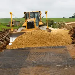 Żółty spychacz Caterpillar przygotowuje teren pod utwardzenie drogi, zasypując wykop piaskiem. Na ziemi rozłożona siatka.