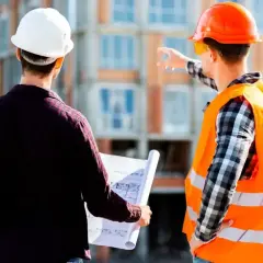 Dwóch budowlańców w kaskach analizuje plany. Jeden wskazuje kierunek, drugi trzyma zgubiony dziennik budowy.
