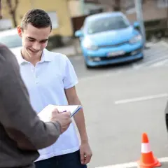 Młody mężczyzna z aparatem na zębach uśmiecha się, gdy instruktor jazdy zapisuje coś na kartce. Niedługo dowie się, od ilu lat można jeździć autem.