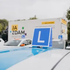 Na dachu białego samochodu nauki jazdy stoi niebieska tablica z literą "L". W tle ciężarówka z napisem "JA ZDAM". Ile można mieć błędów na egzaminie teoretycznym?
