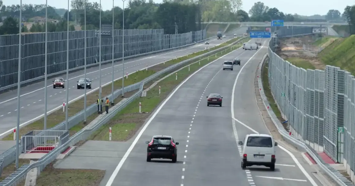Samochody poruszają się lewym pasem w terenie zabudowanym, mijając ekrany akustyczne.