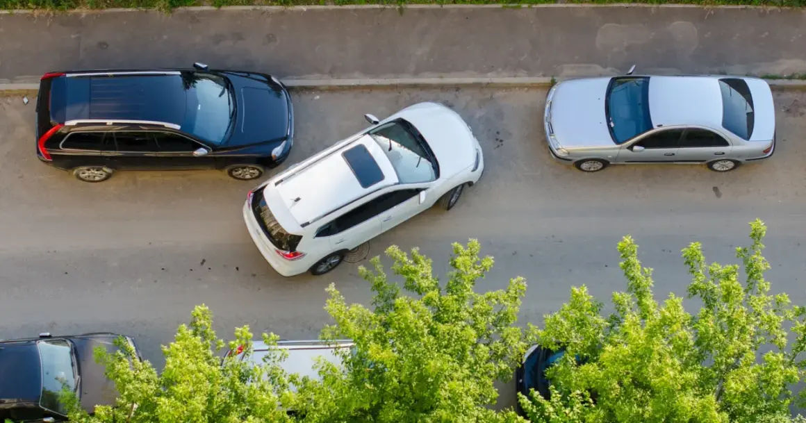 Biały SUV wykonuje parkowanie równoległe przodem między czarnym SUV-em a srebrnym sedanem.