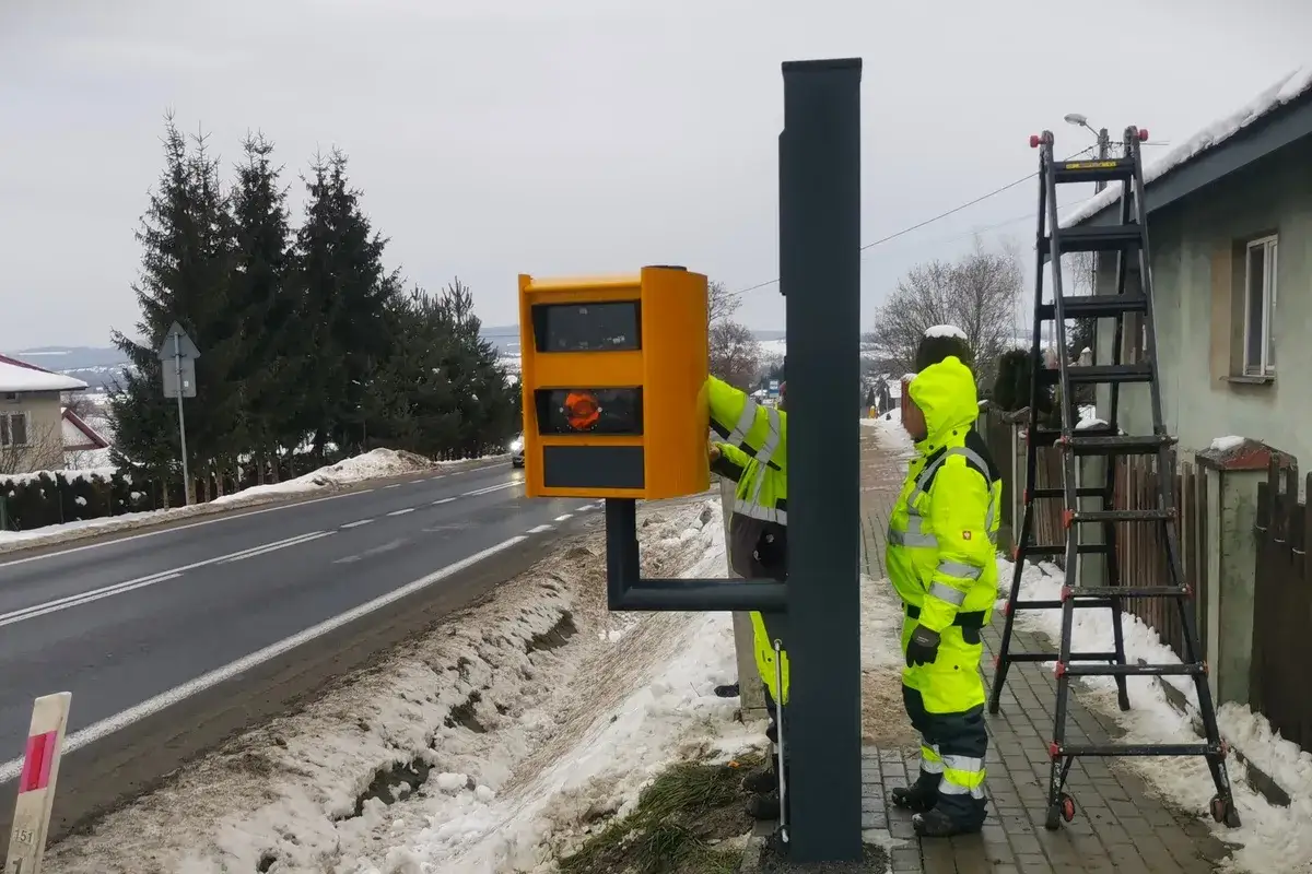 Montaż fotoradarów w Polsce. Dwóch pracowników w kamizelkach odblaskowych instaluje żółty fotoradar przy drodze.