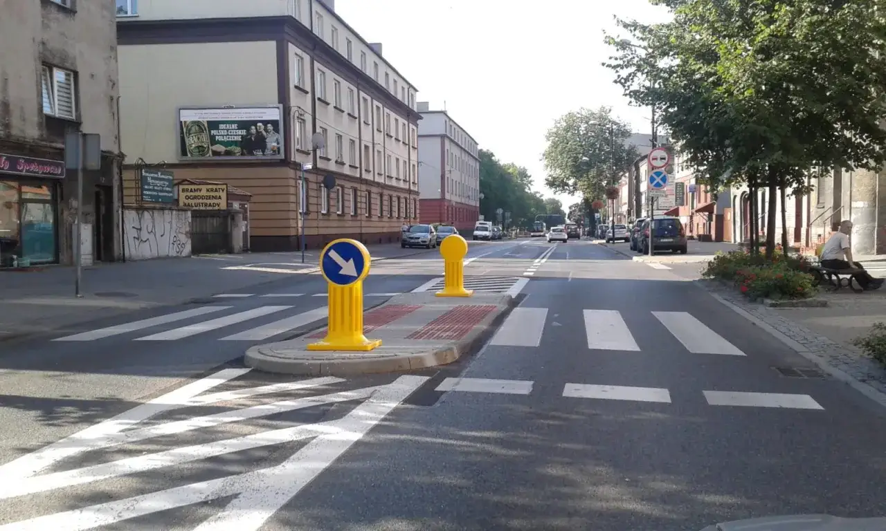 Przejście dla pieszych z wysepką, z żółtymi słupkami i niebieskimi strzałkami, ułatwia bezpieczne przekraczanie ulicy.