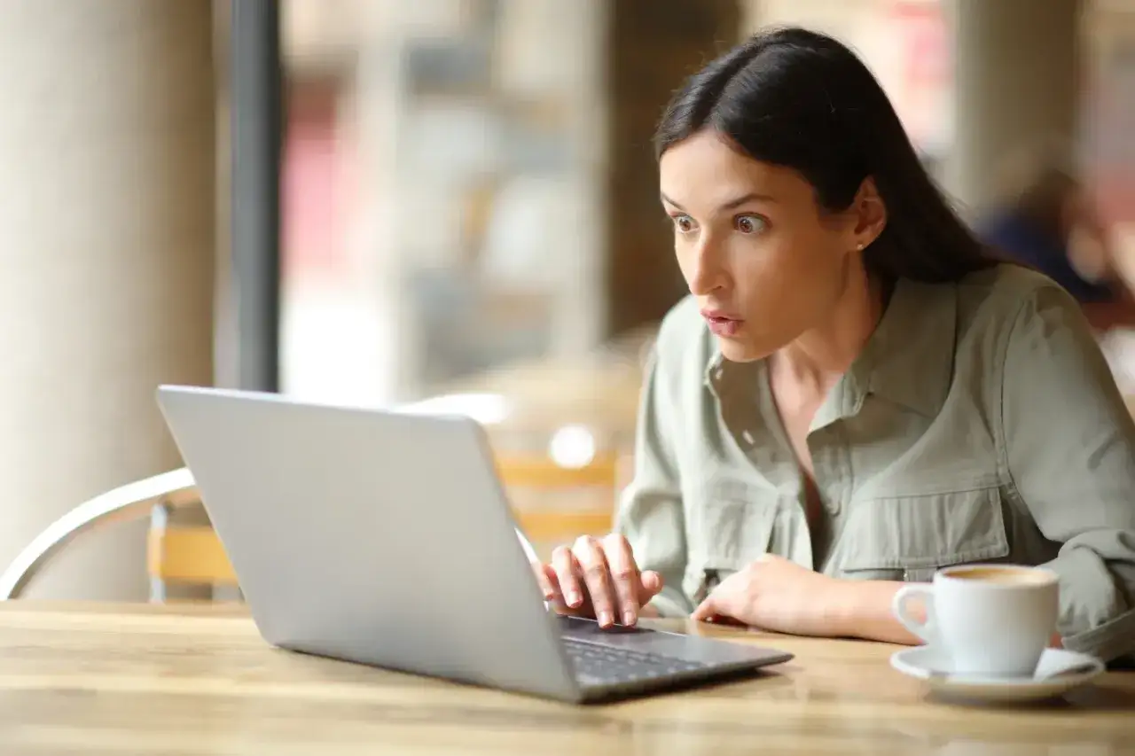 Zaskoczona kobieta przy kawie patrzy na ekran laptopa, szukając odwołania do kio.