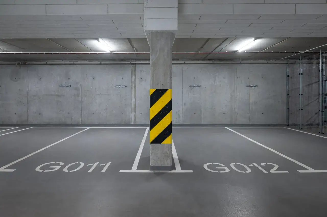 Puste miejsce postojowe G011 i G012 w podziemnym parkingu. Betonowe ściany i sufit, oświetlenie jarzeniowe.